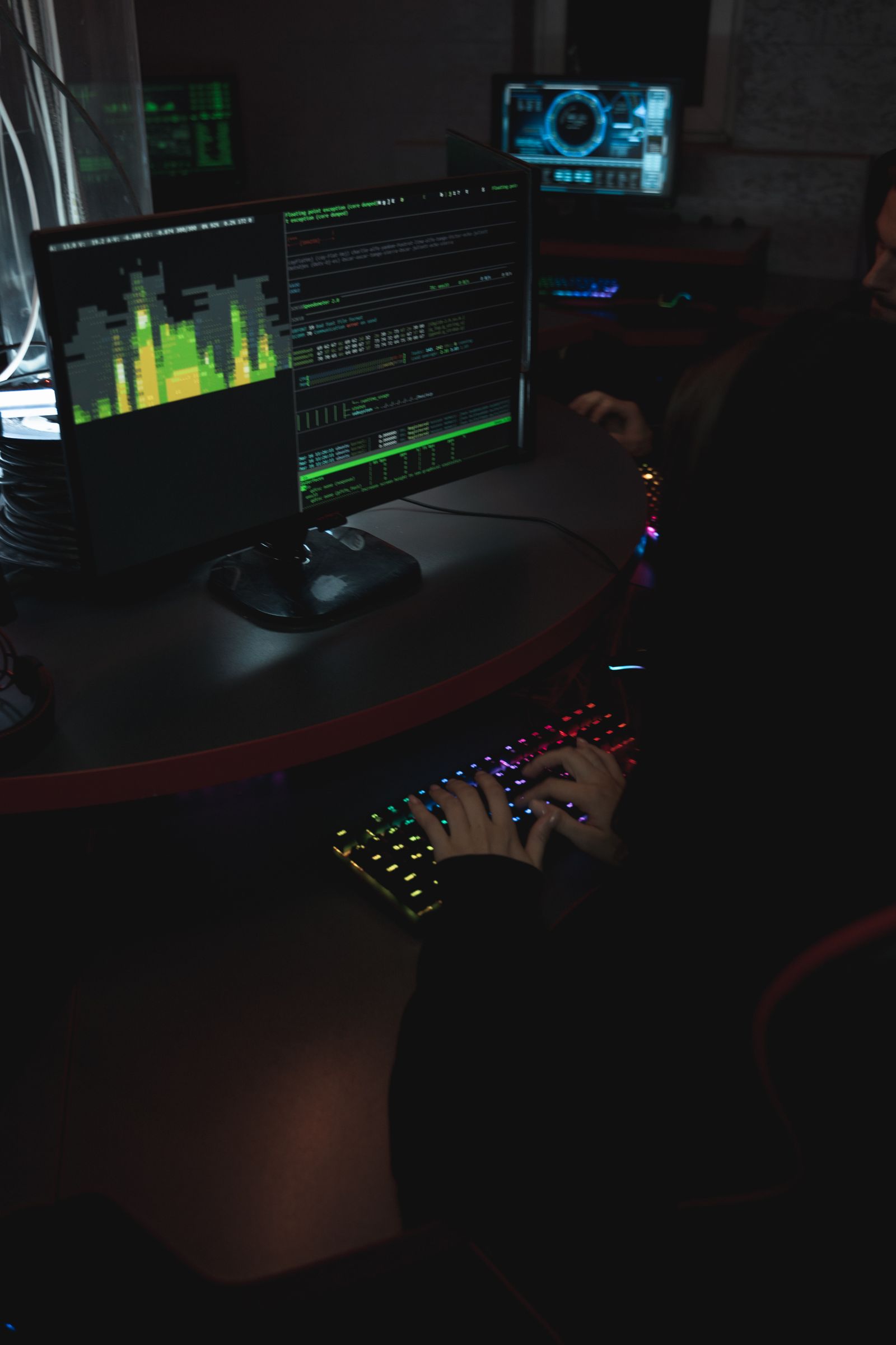 Pessoa digita em um teclado retroiluminado, em uma sala escura, diante de monitores com gráficos de terminal