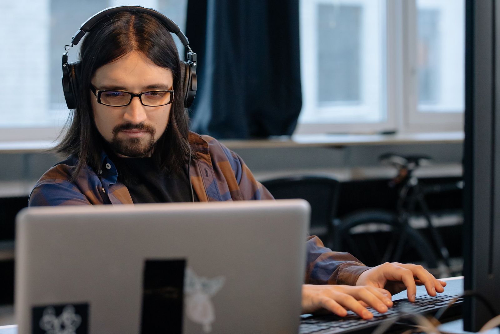 Homem de cabelo comprido, com fones de ouvido e óculos, digita em um notebook no escritório