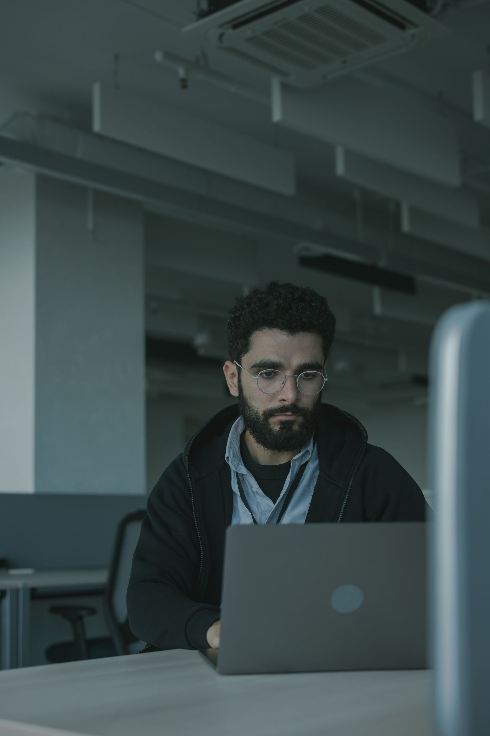 Homem de barba e óculos trabalha em um notebook em um escritório amplo e vazio