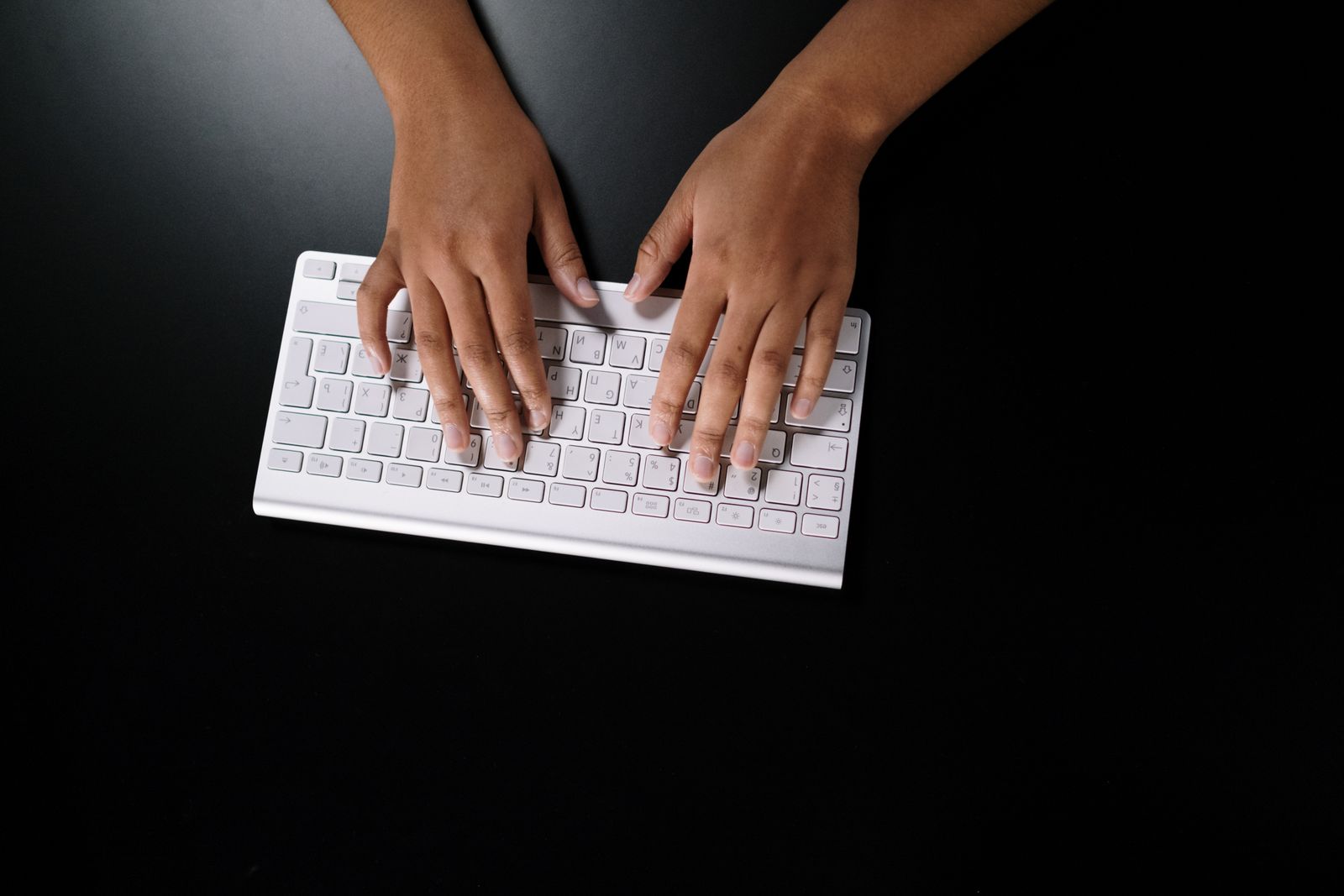 Vista de cima das mãos de uma mulher digitando em um teclado branco sobre uma mesa preta