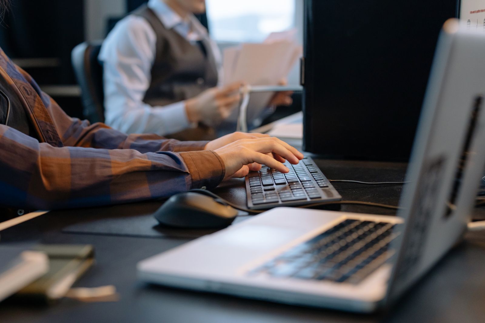 Mãos digitam em um teclado em uma mesa de escritório, com um notebook à frente e um colega segurando papéis ao fundo