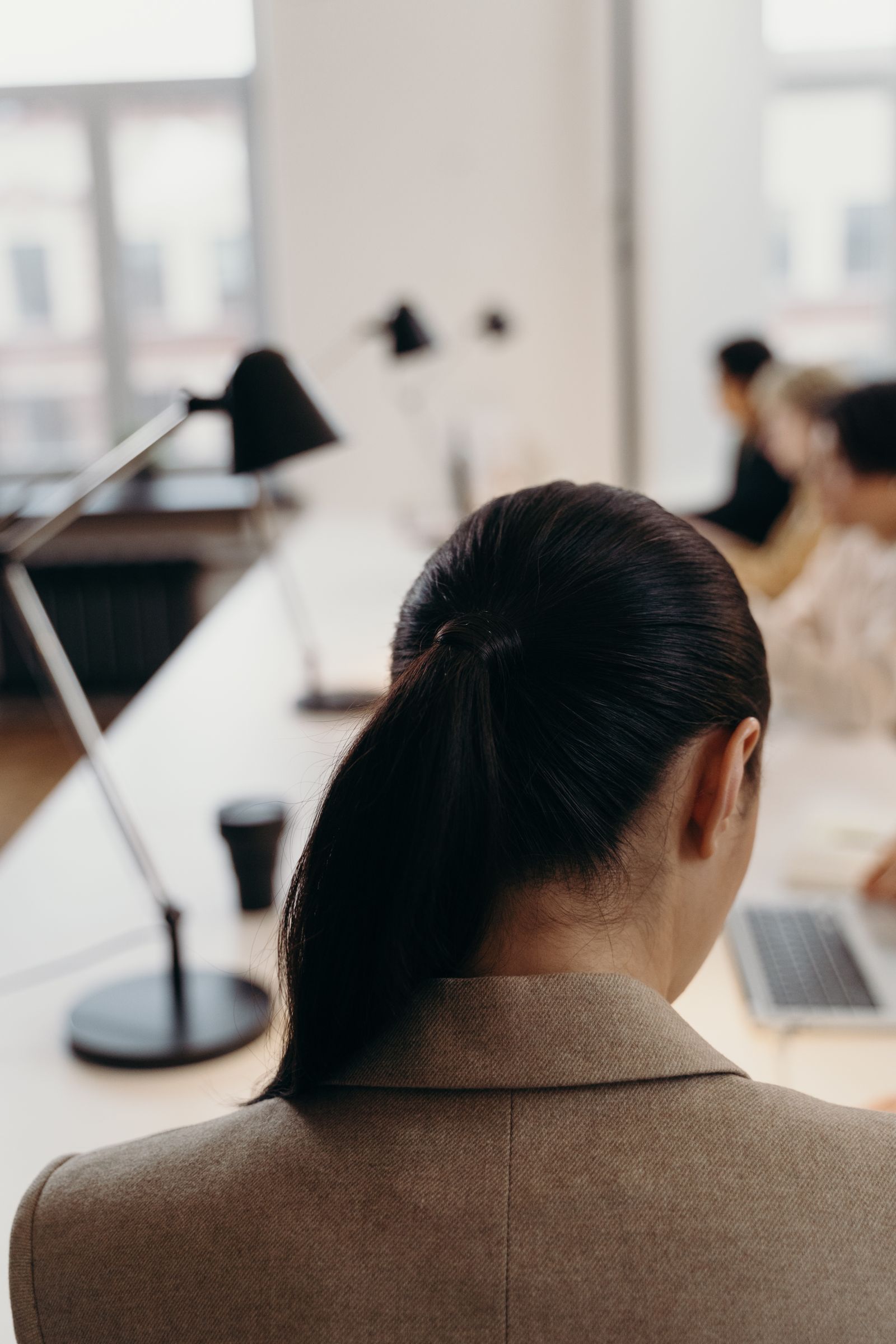 Mulher de rabo de cavalo, vista de costas, em um escritório com luminárias de mesa e colegas desfocados trabalhando em notebooks